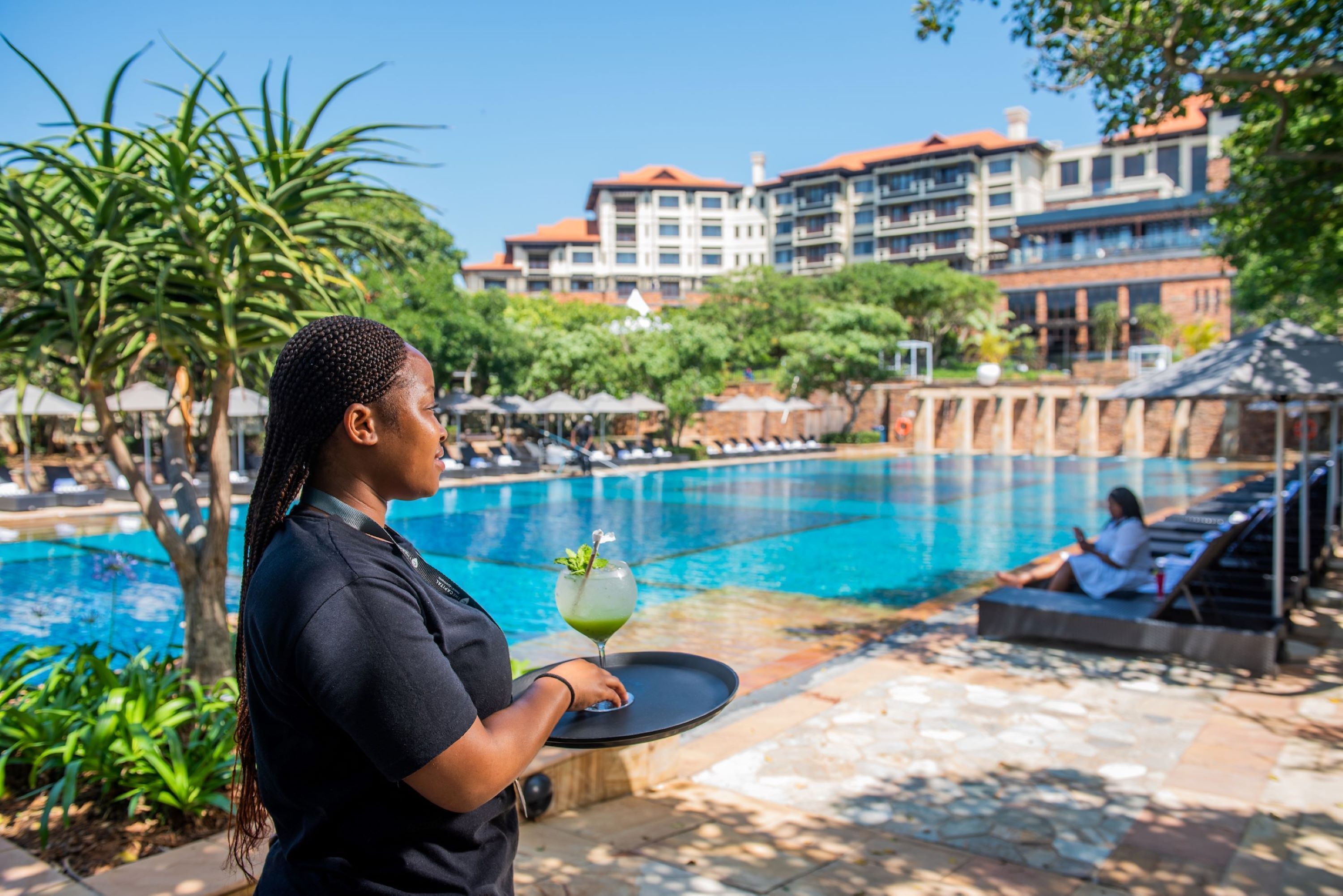 A poolside area with a person holding a tray, a pool, and a building with balconies in the background.