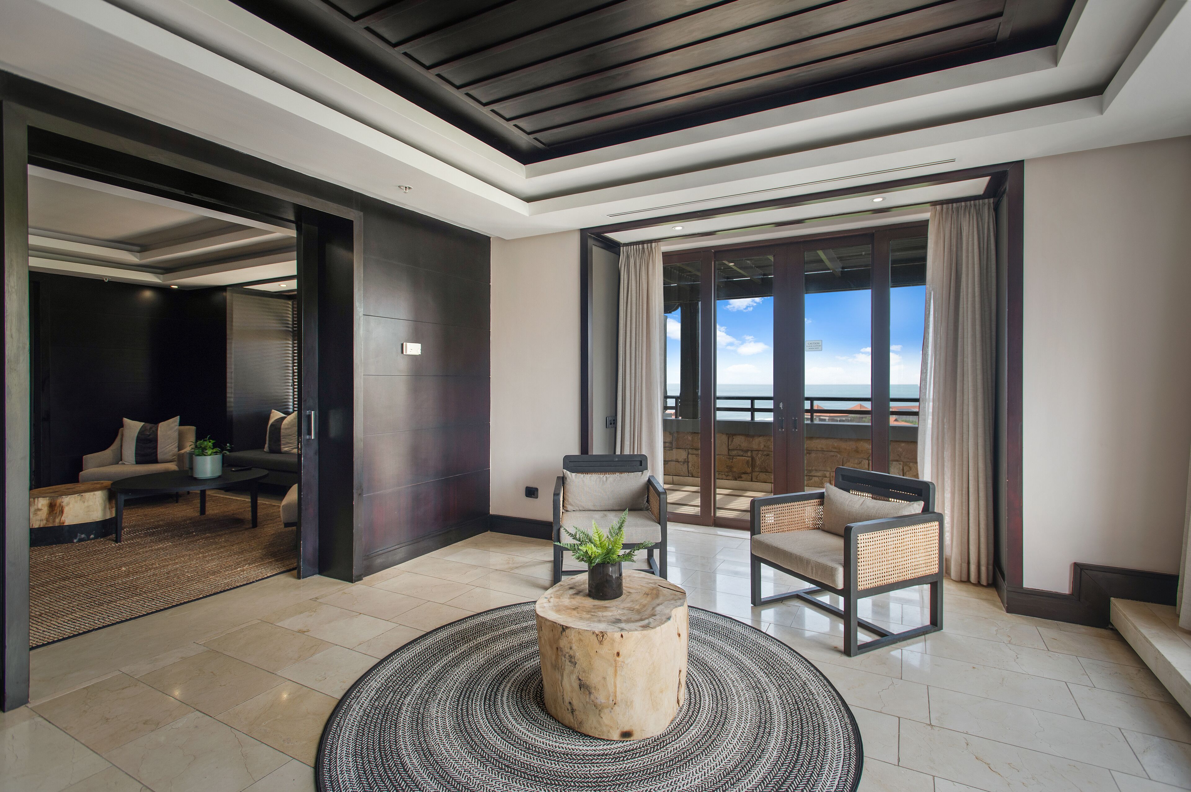 Quarto de hotel moderno com uma varanda grande, área de estar com sofá e cadeiras, e uma mesa redonda de madeira com uma planta.