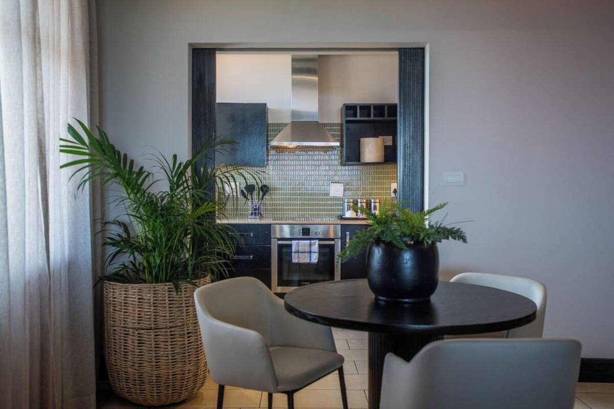 A dining area with a round table, two chairs, a large potted plant, and a view into a kitchen with a tiled backsplash and modern appliances.