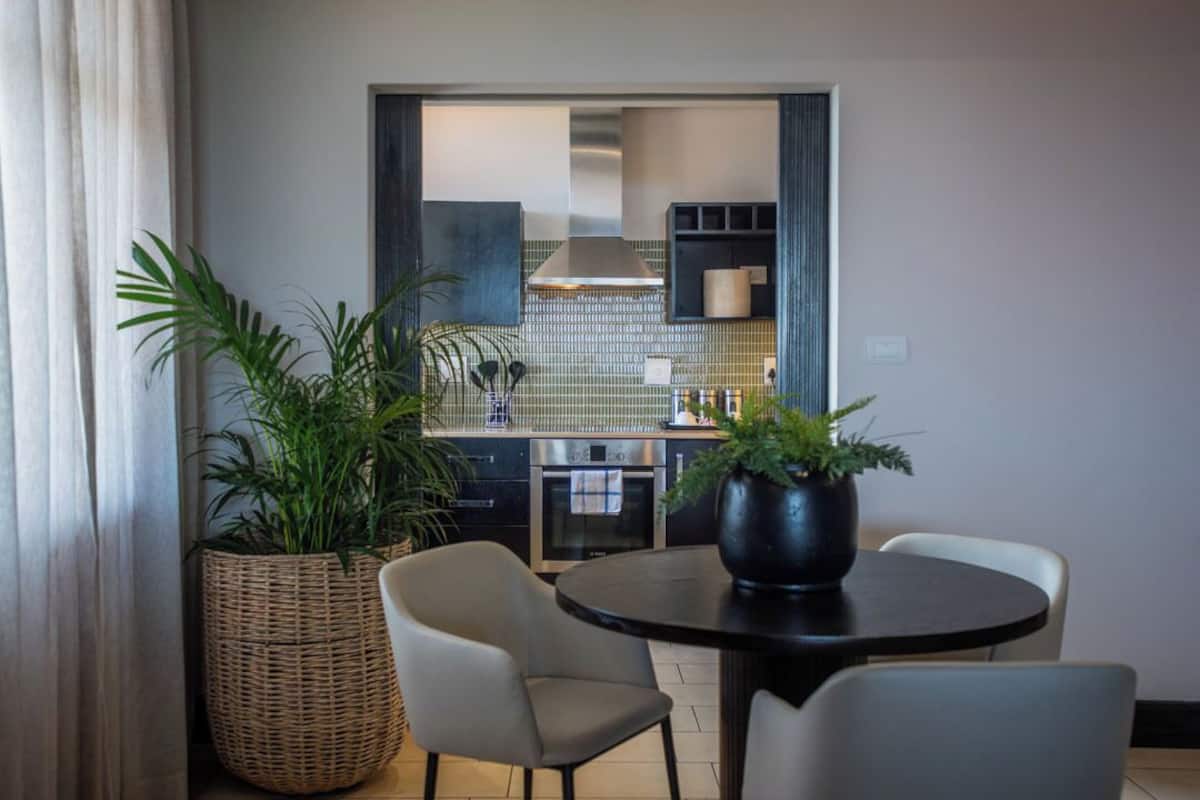 A dining area with a round table, two chairs, a large potted plant, and a view into a kitchen with a tiled backsplash and modern appliances.