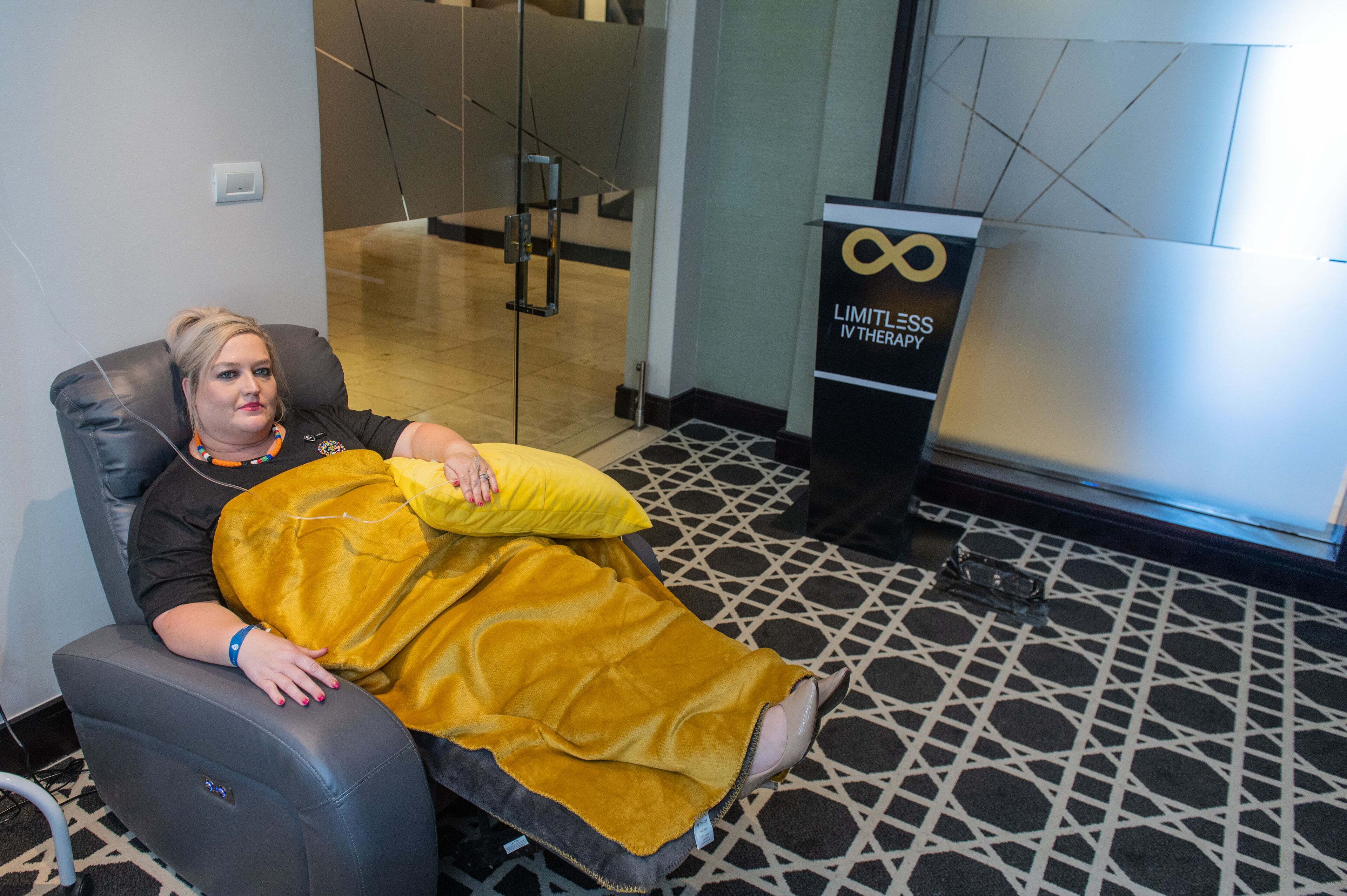 A person reclining in a massage chair with a yellow blanket, in a room with a patterned floor and a glass door leading to another area.