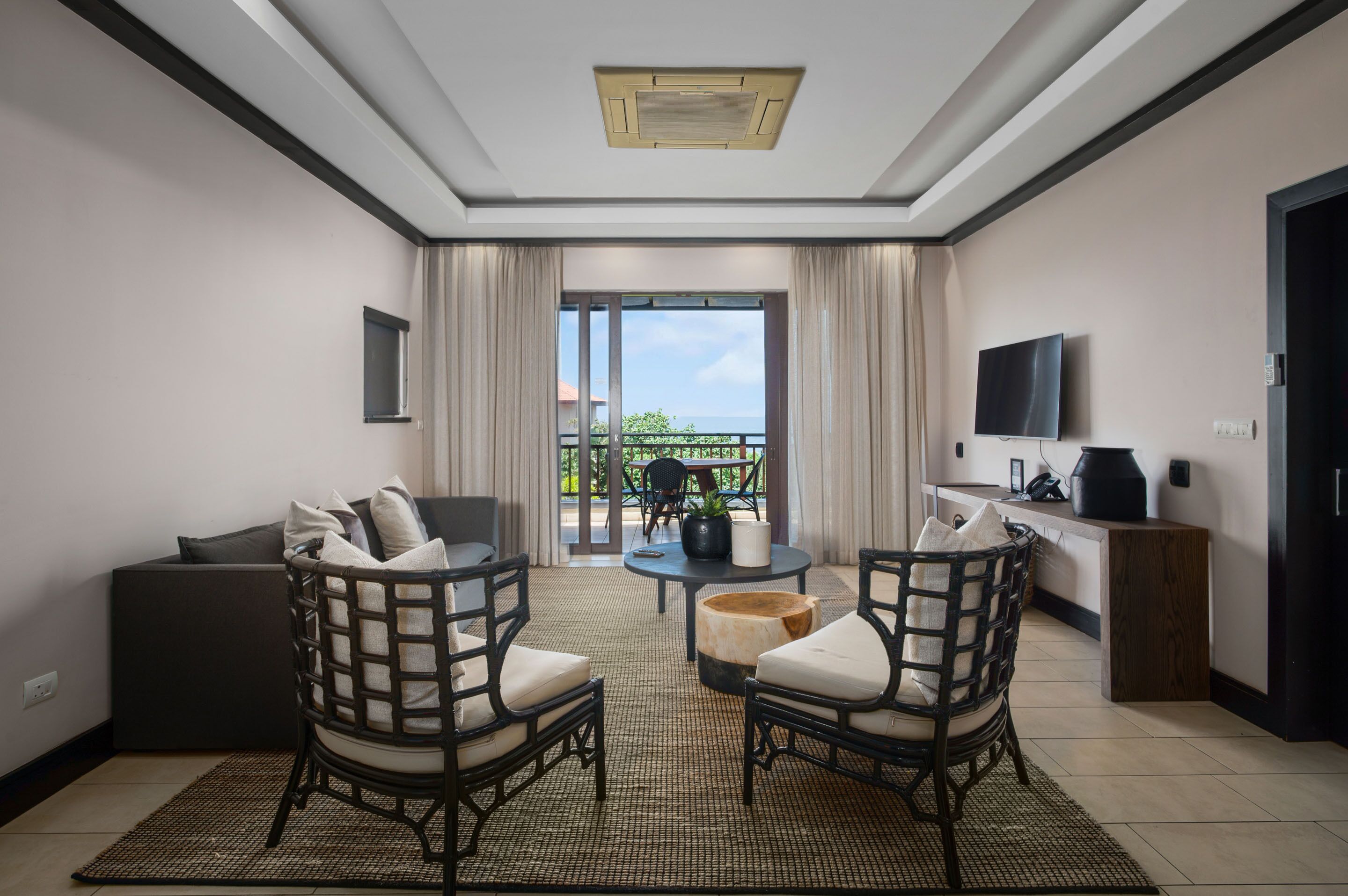A modern living room with a sofa, two armchairs, a coffee table, and a TV. There is a balcony with a view of the outdoors.
