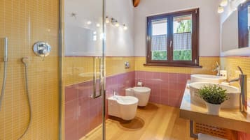 A bathroom with a glass shower enclosure, two toilets, a sink, and a window.