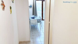 Un couloir étroit mène à une salle à manger où se trouvent une table et des chaises, une fenêtre avec des rideaux et une porte.