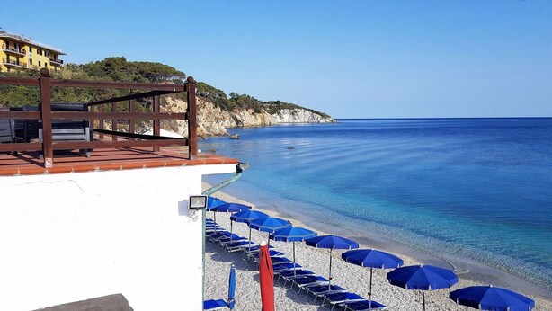 Une plage à l’eau d’un bleu limpide, une terrasse en bois avec des sièges et des parasols.