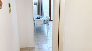 Un couloir étroit mène à une salle à manger où se trouvent une table et des chaises, une fenêtre avec des rideaux et une porte.