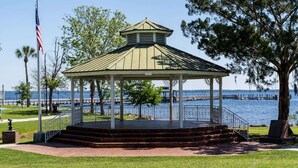 Un kiosque doté d’un toit en métal et de colonnes blanches, situé près d’une zone riveraine avec un quai et des arbres.