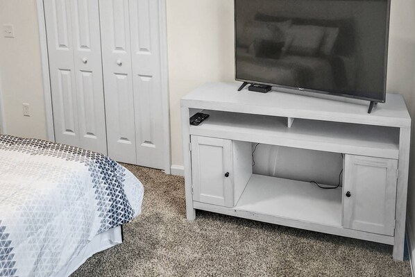 A bedroom with a bed, a white TV stand with a flat-screen TV, and a white wardrobe.
