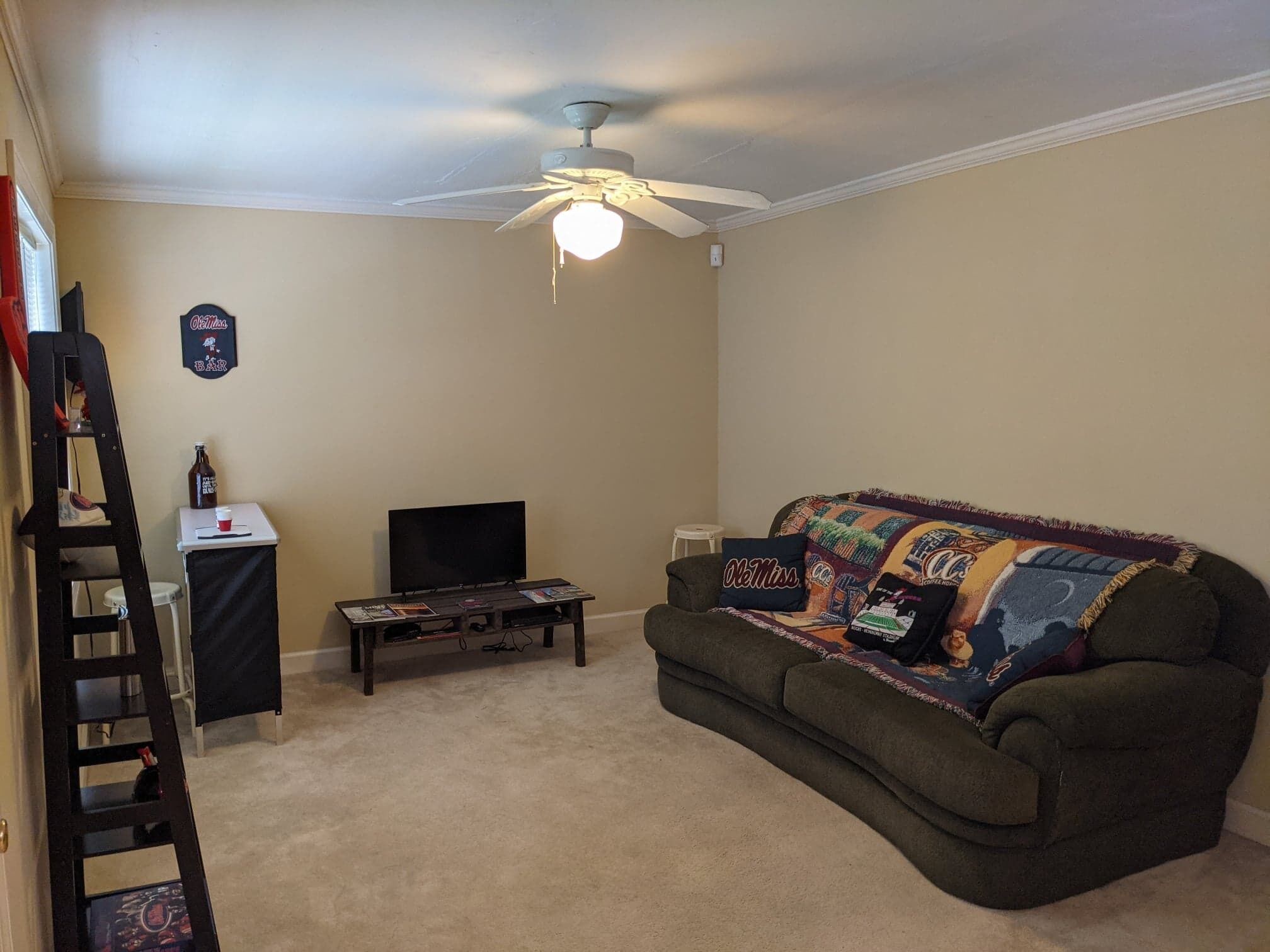 A living room with a ceiling fan, a television on a stand, a couch with a colorful blanket, and a small table with a bottle and a cup.