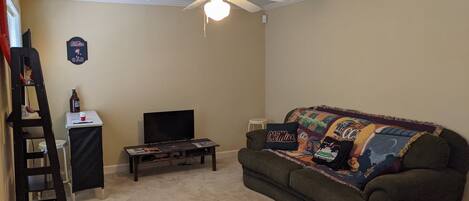 A living room with a ceiling fan, a television on a stand, a couch with a colorful blanket, and a small table with a bottle and a cup.