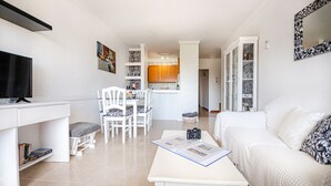 A modern living room with a white sofa, a dining area with white chairs, a kitchen in the background, and a TV on a white desk. - 2 bedroom apartment 200 m. from the beach (Alcúdia)