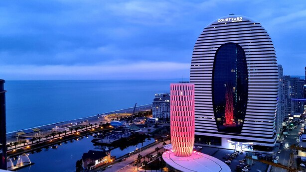 A modern hotel with a distinctive arched entrance and a large central window, situated in a cityscape with a view of the sea. - Orbi City Apartment (Batumi)