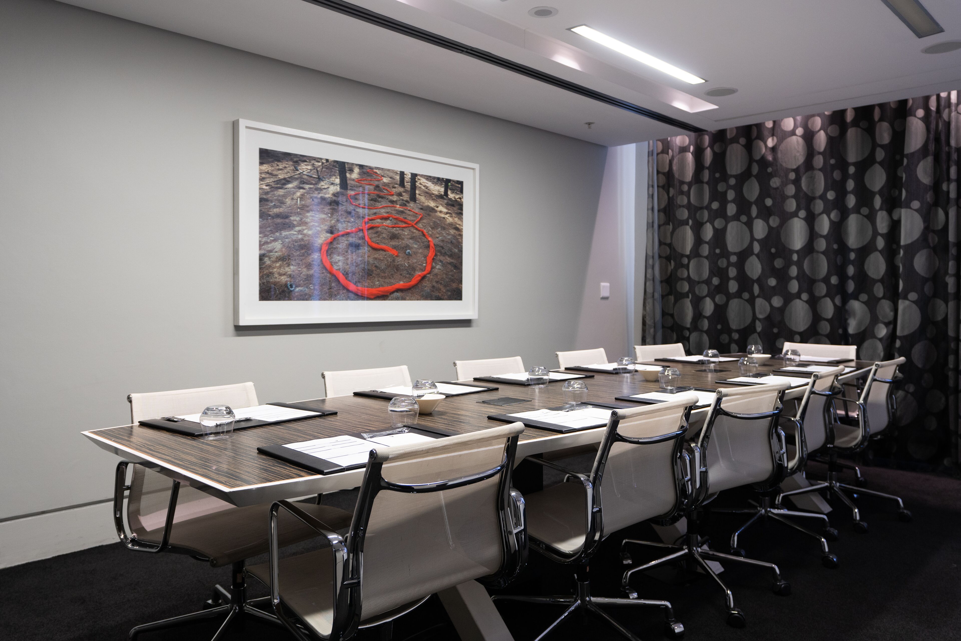 A conference room with a long table, chairs, a framed abstract painting, and a patterned curtain.