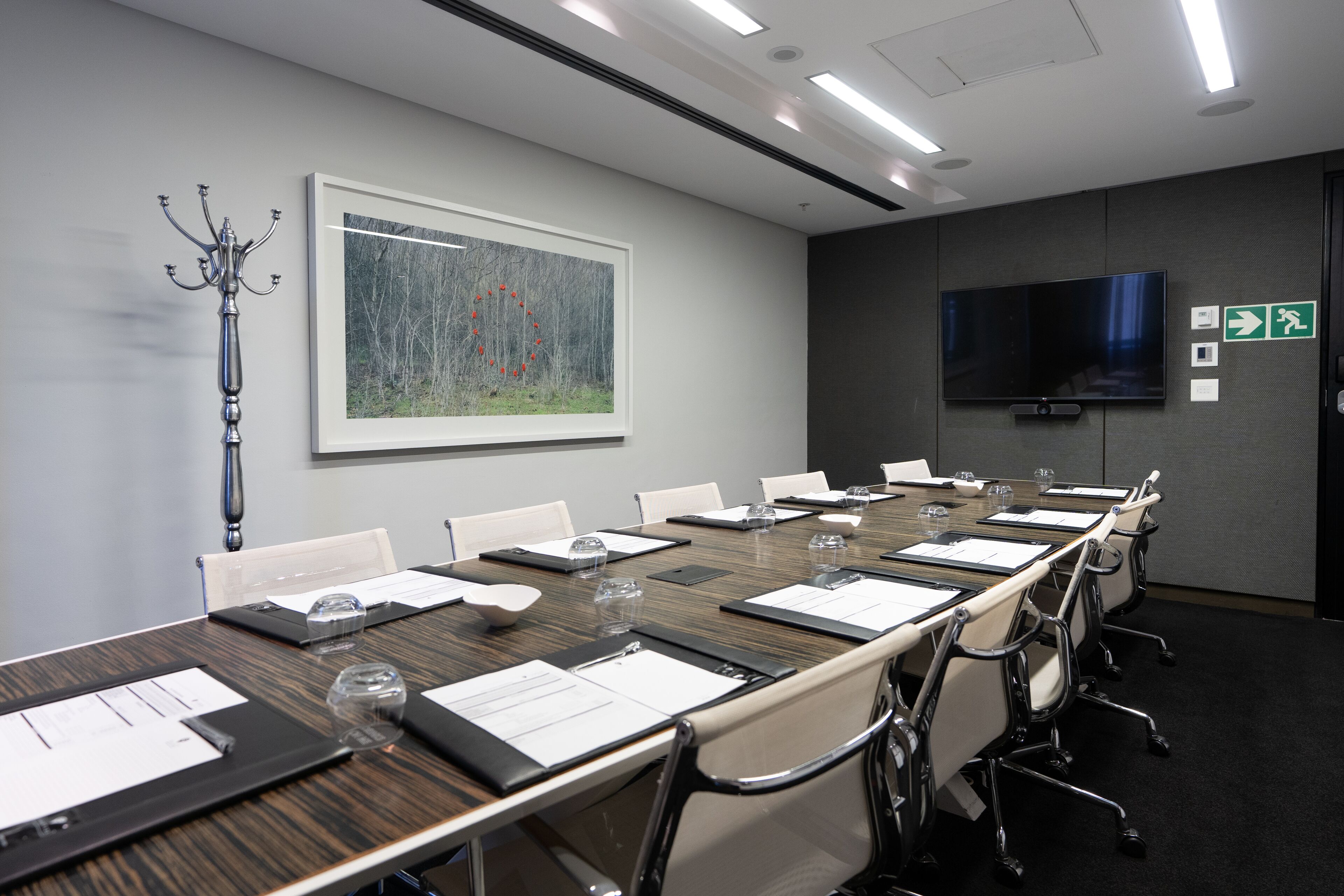 Uma sala de conferências com uma mesa comprida, cadeiras, tela de projetor, um quadro e um cabideiro.