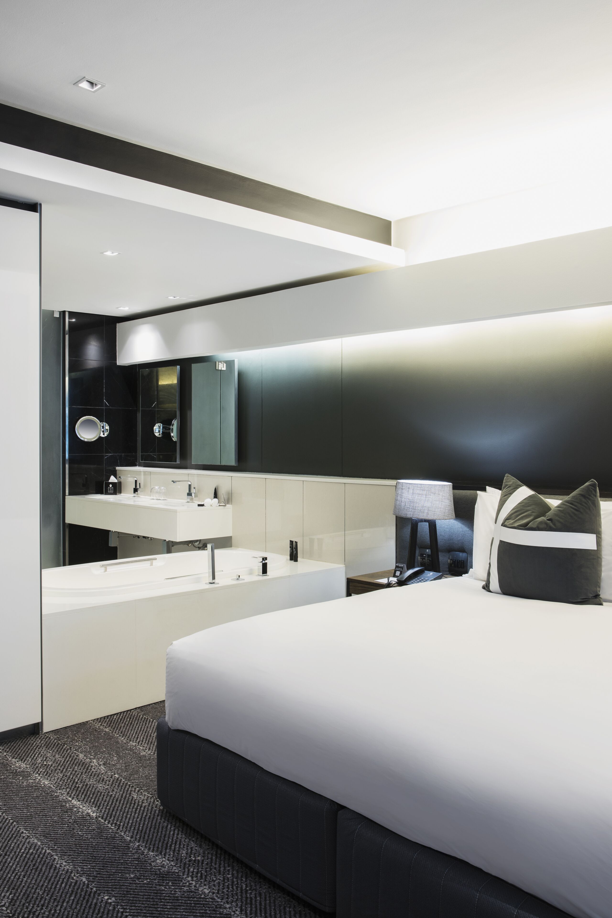 A modern hotel room with a large bed, a built-in vanity with a mirror, and a bedside table with a lamp.