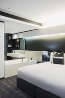 A modern hotel room with a large bed, a built-in vanity with a mirror, and a bedside table with a lamp.