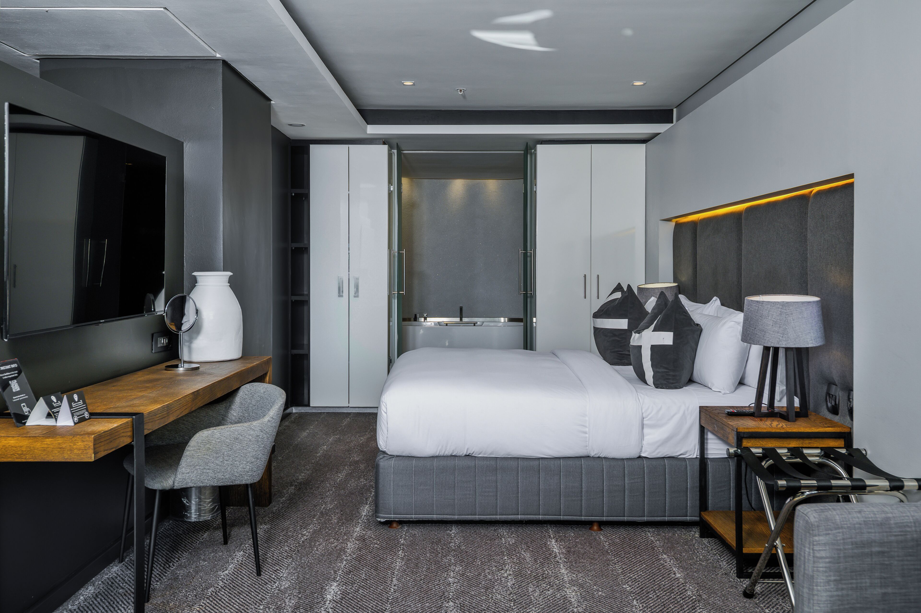 Una habitación de hotel moderna con cama, escritorio, silla, televisor, lámpara y armario.