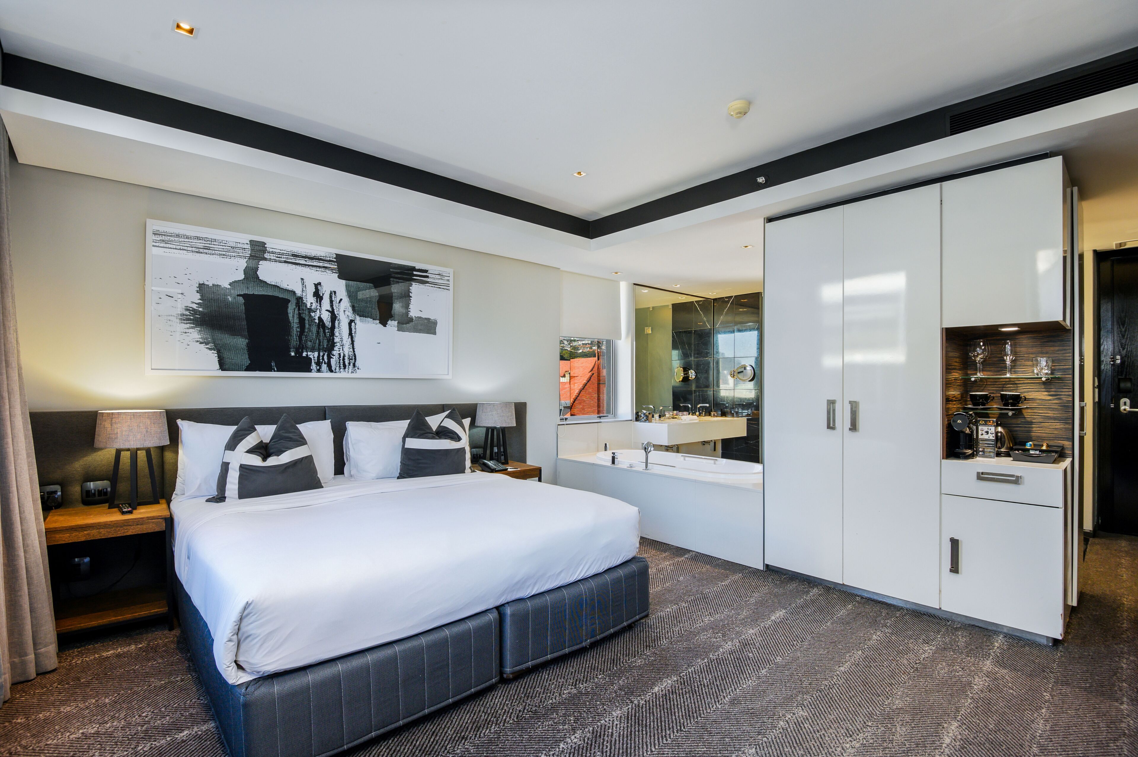 Ein modernes Hotelzimmer mit einem großen Bett, einem Nachttisch mit Lampe, einem großen Spiegel, einem Kleiderschrank und einem Badezimmer mit Waschbecken und Spiegel.