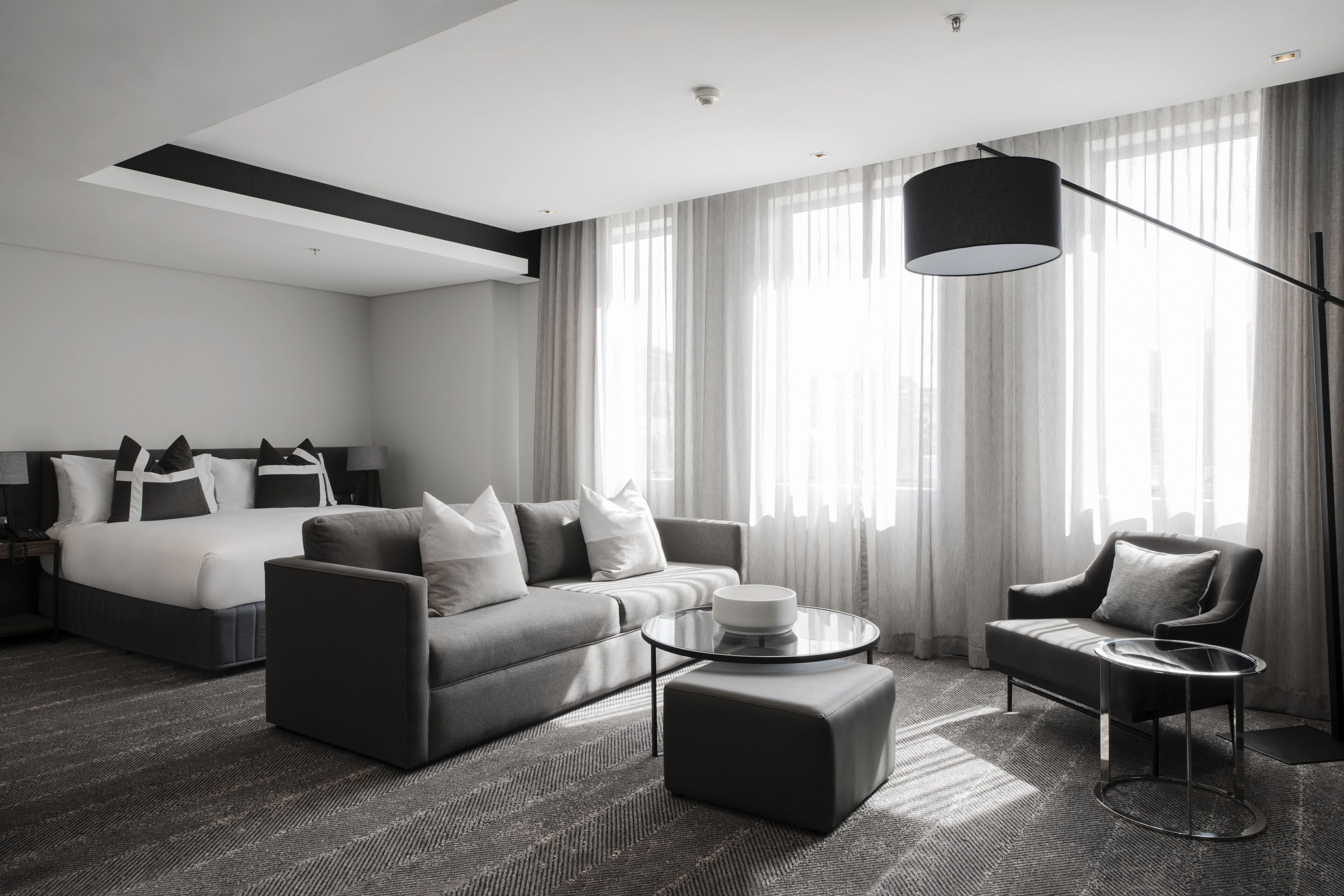 A modern hotel room with a bed, sofa, armchair, and a round coffee table.