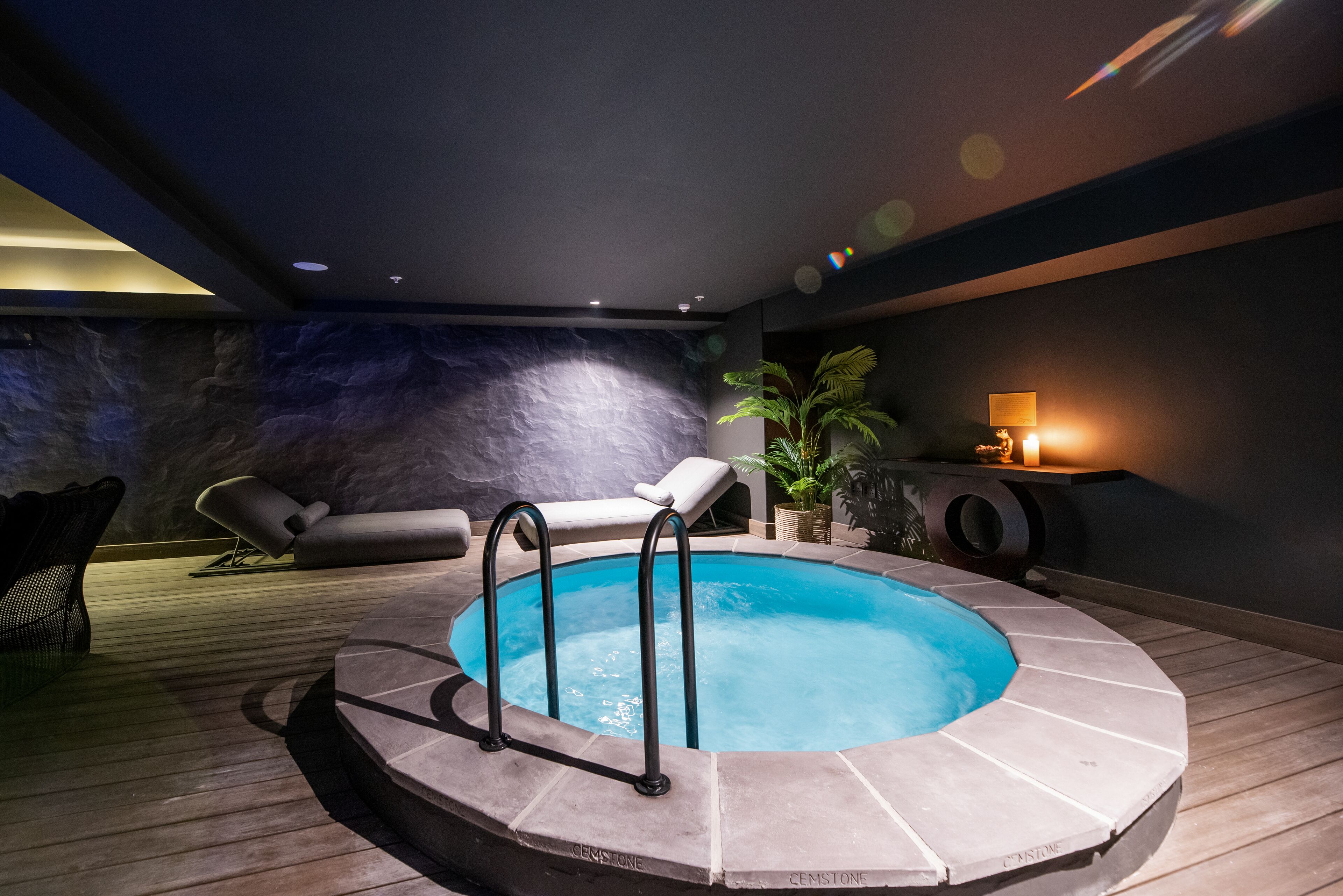 A modern indoor swimming pool with a stone edge, lounge chairs, and a wall with a textured finish.