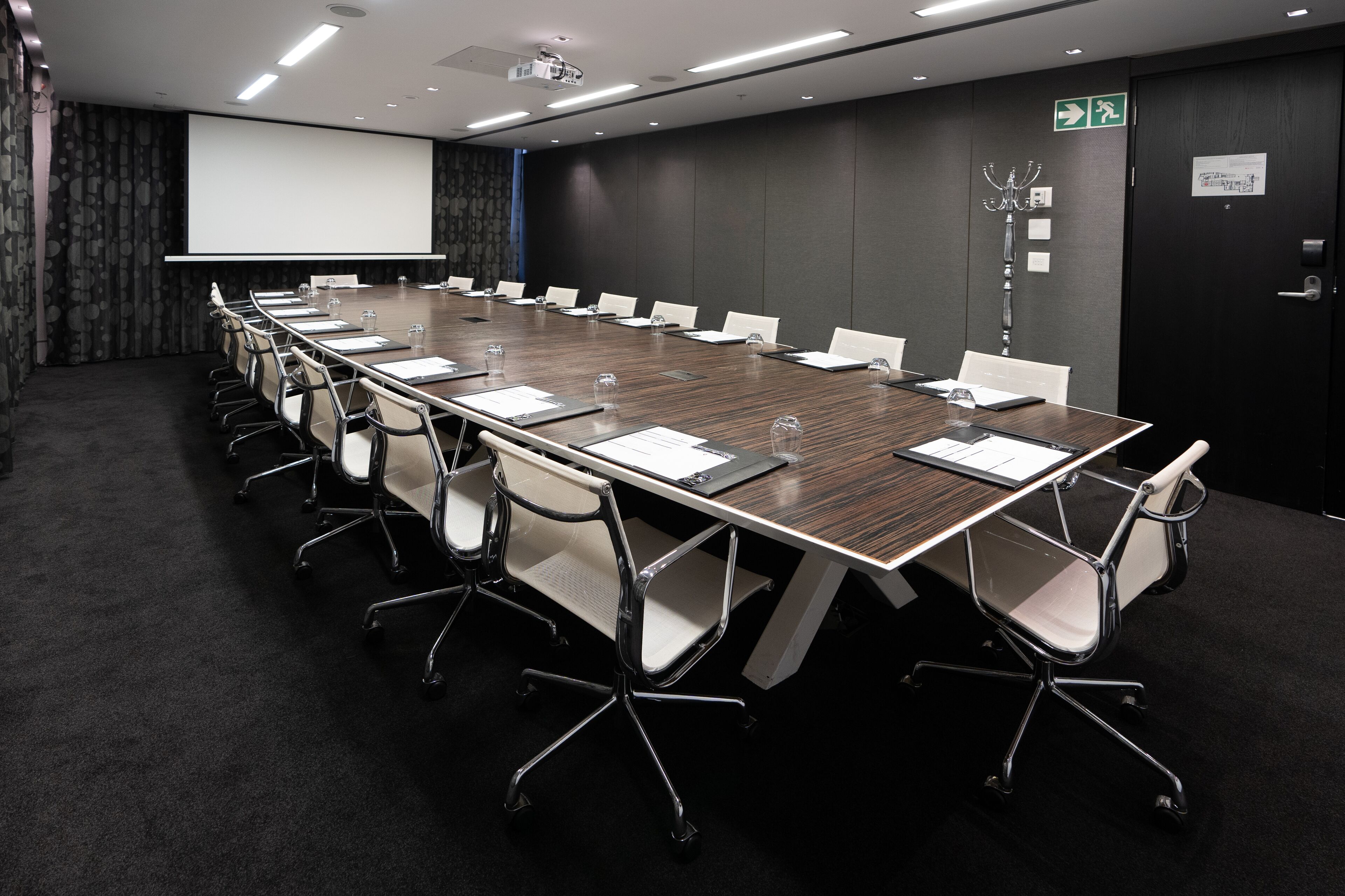 A conference room with a long table, white chairs, a projector screen, and a dark carpeted floor.
