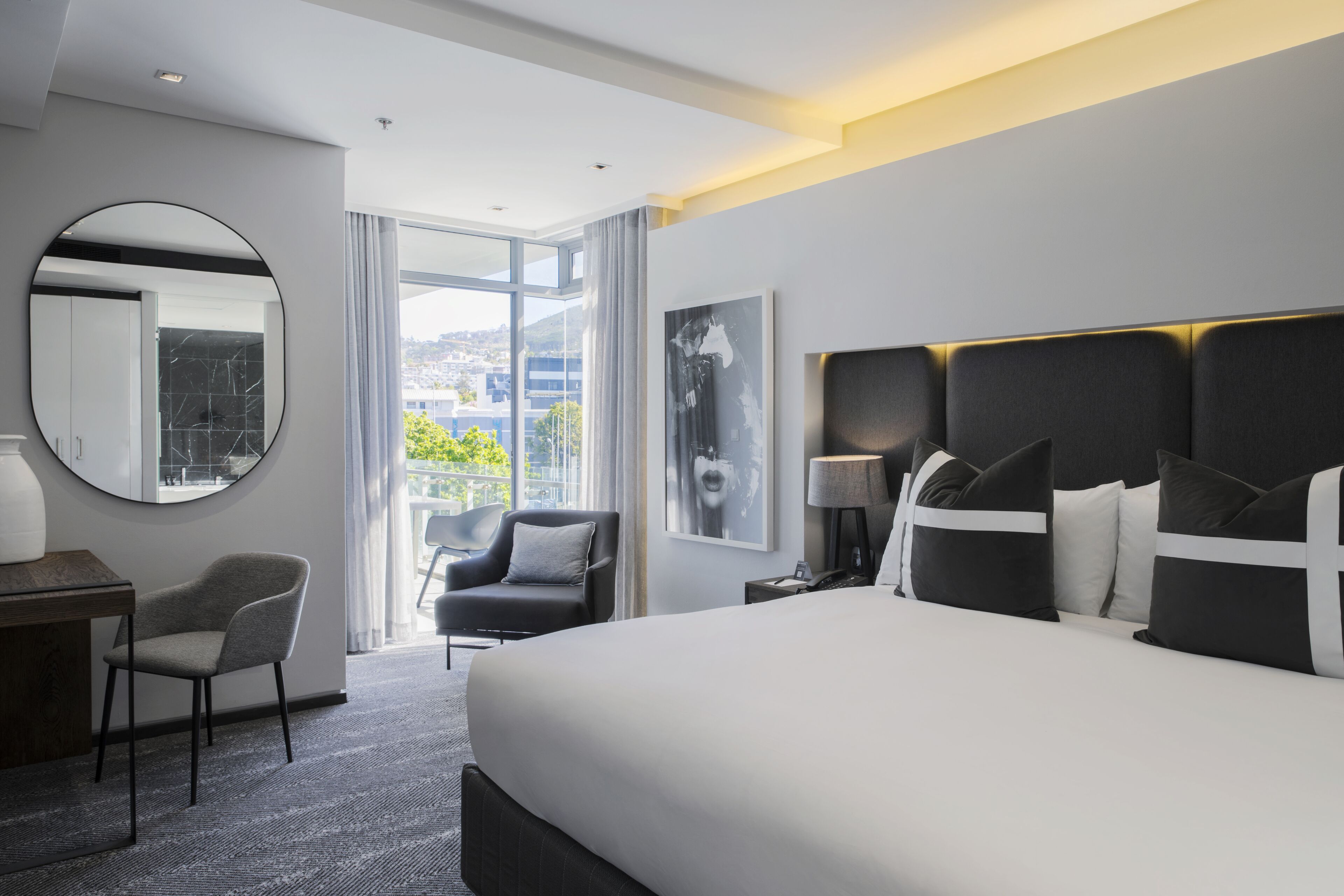 A modern hotel room with a large bed, a round mirror, a desk, a chair, and a view of the city.