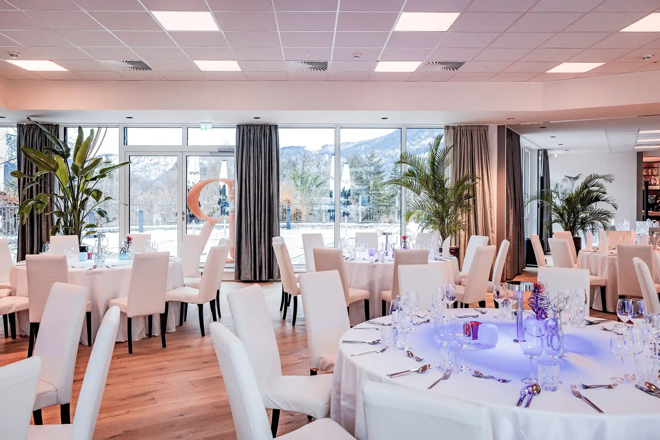 A spacious dining area with round tables set for a formal event, large windows offering a view of the outdoors, and a modern ceiling design.