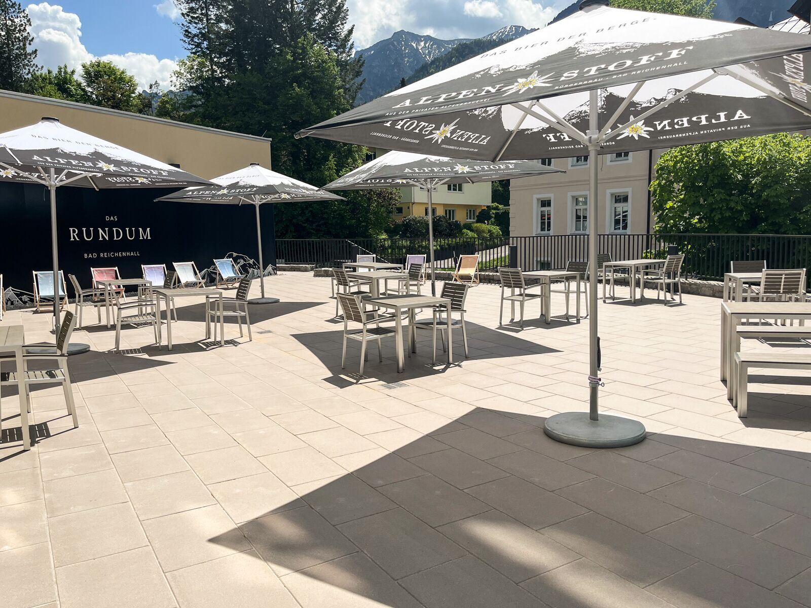 An outdoor seating area with tables and chairs under umbrellas, a building with 'ALPEN STOFF' branding, and a sign for 'RUNDUM BAD REICHENHALL'.