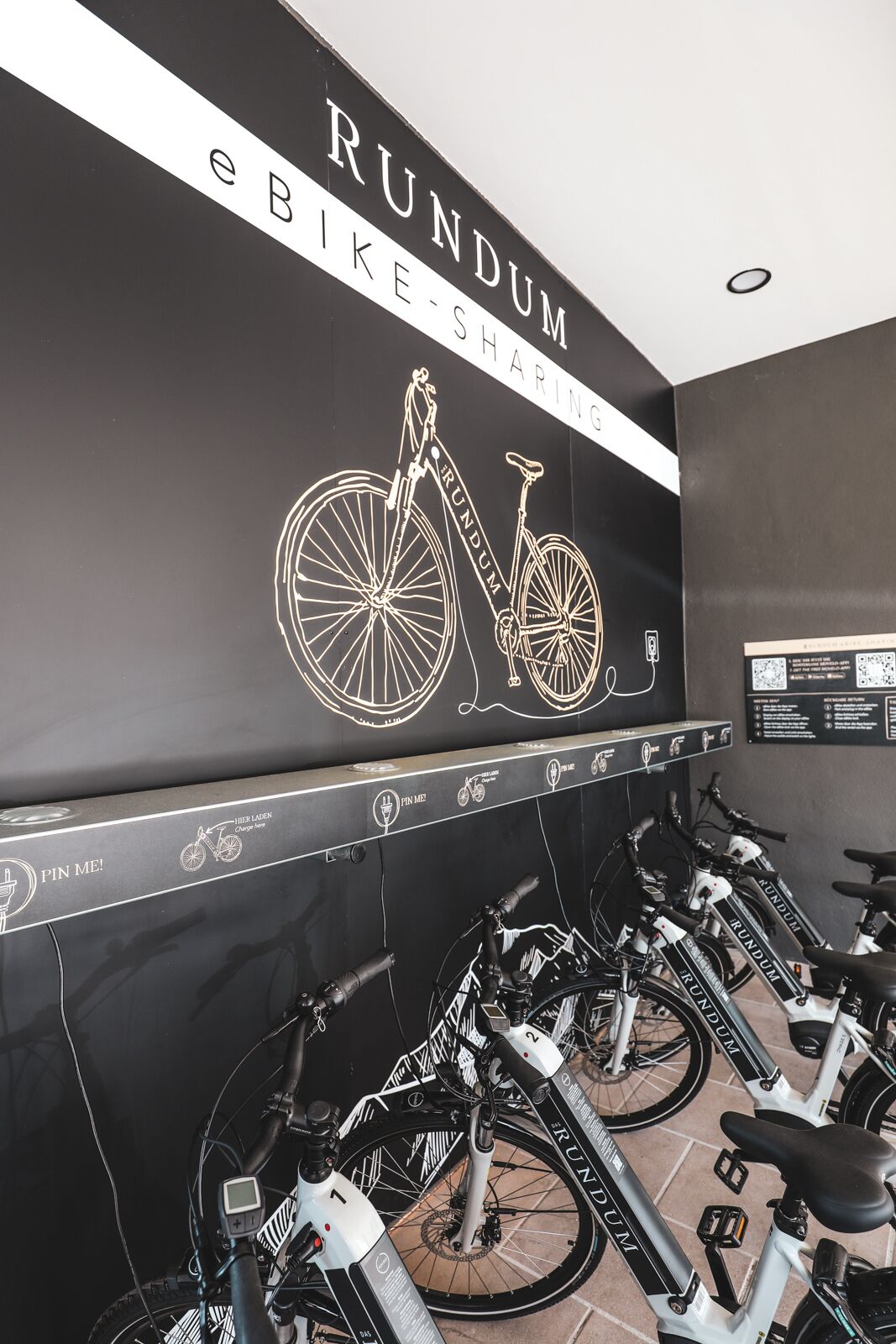 Een deelautofietspunt met meerdere fietsen en een bord dat aan de muur is bevestigd, waarop staat geschreven: 'RUNDUM eBIKE SHARING'.