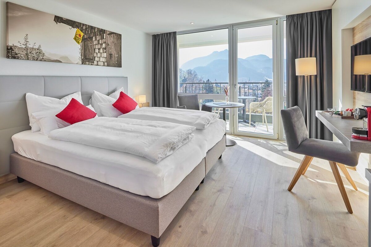 Habitación de hotel con una cama grande, un escritorio, una silla, una mesita y vistas a las montañas.
