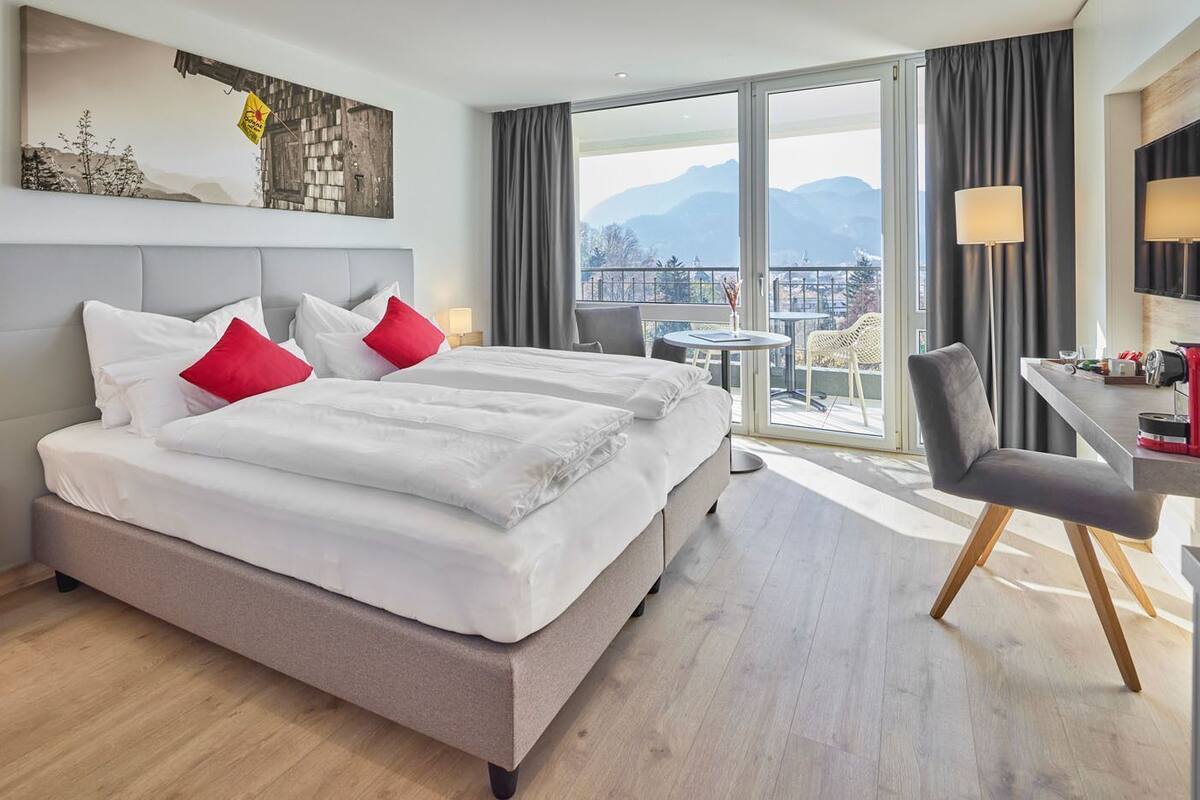 A hotel room with a large bed, a desk, a chair, a small table, and a view of mountains.