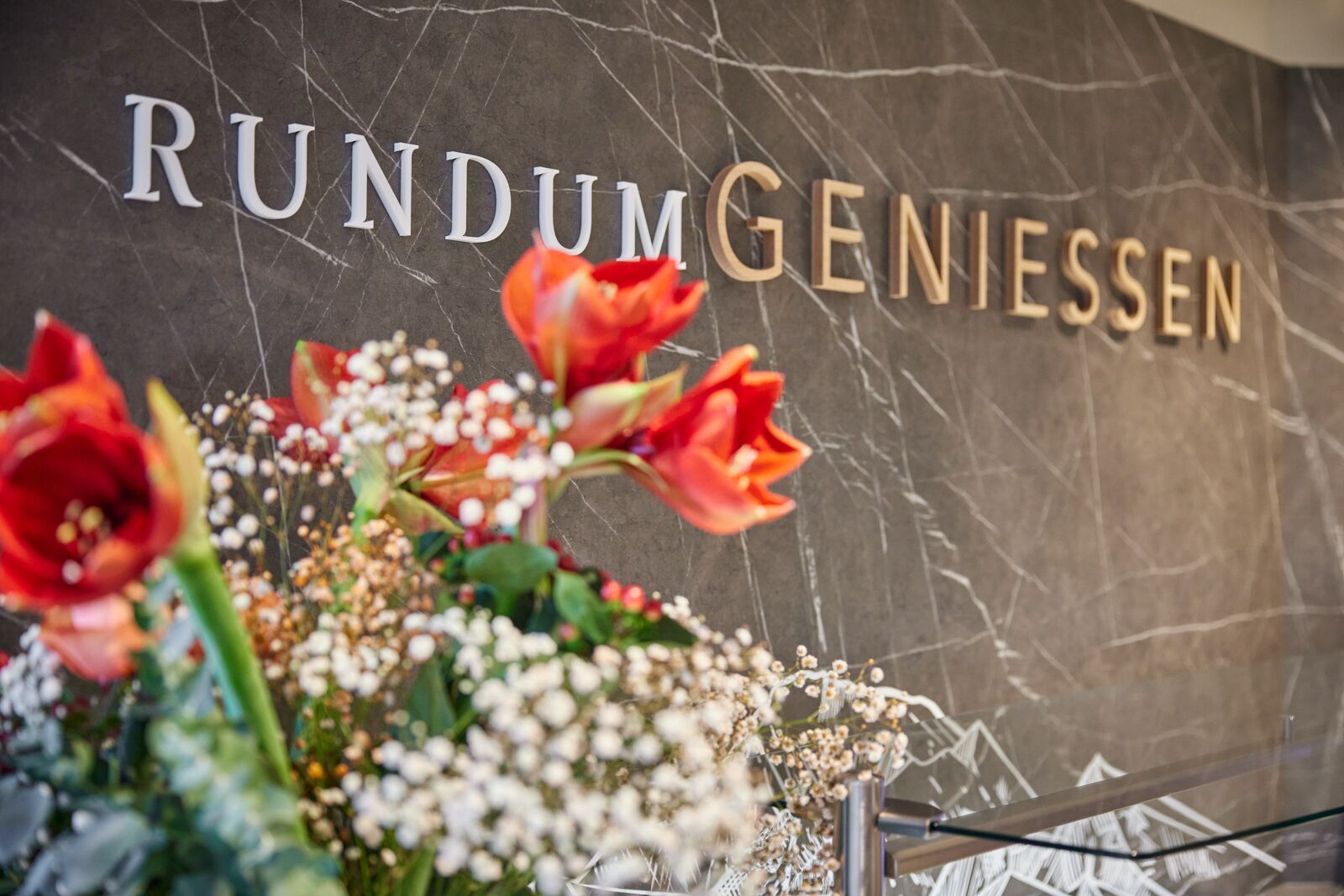 A marble wall with the text "RUNDUM GENIESSEN" and a floral arrangement with red tulips and white flowers.