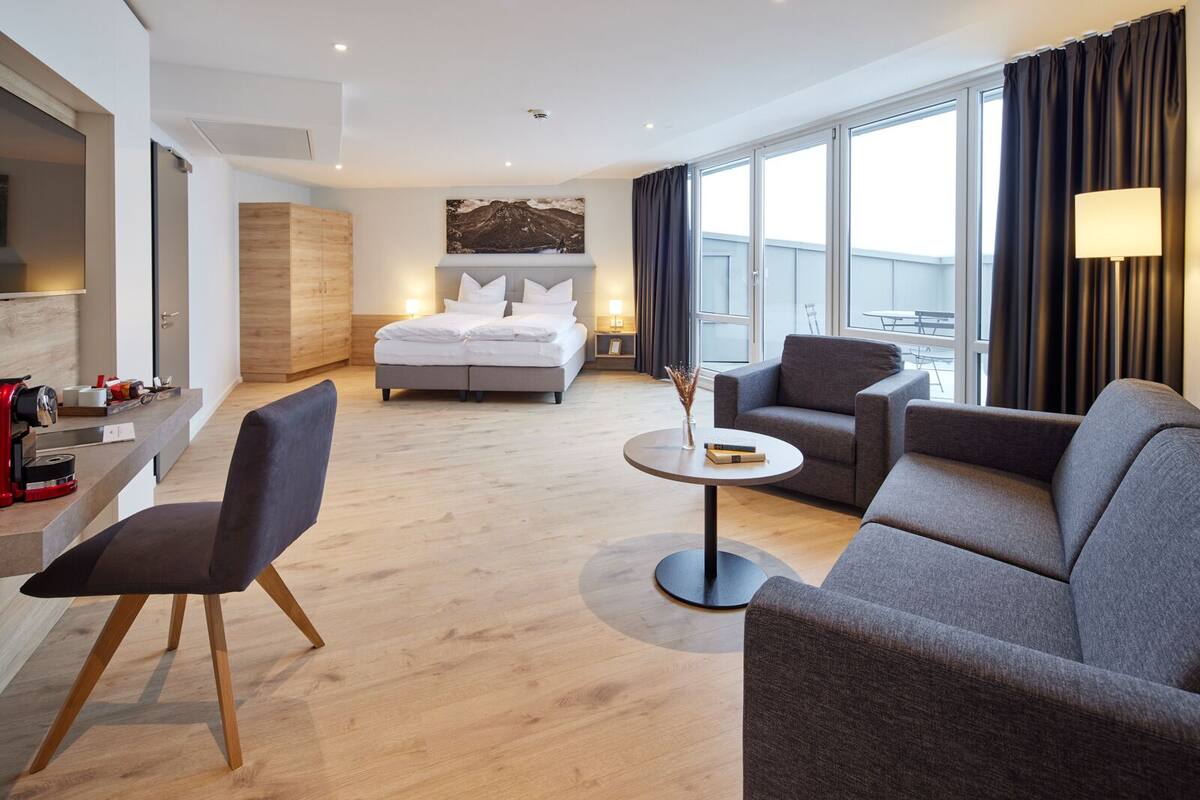 A modern hotel room with a bed, two armchairs, a small table, and a view of the outdoors.
