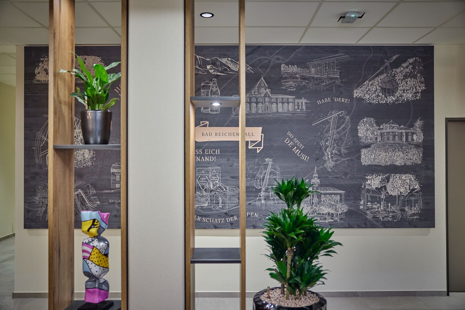 A lobby with a large chalkboard wall displaying architectural drawings, a wooden partition, and two potted plants.