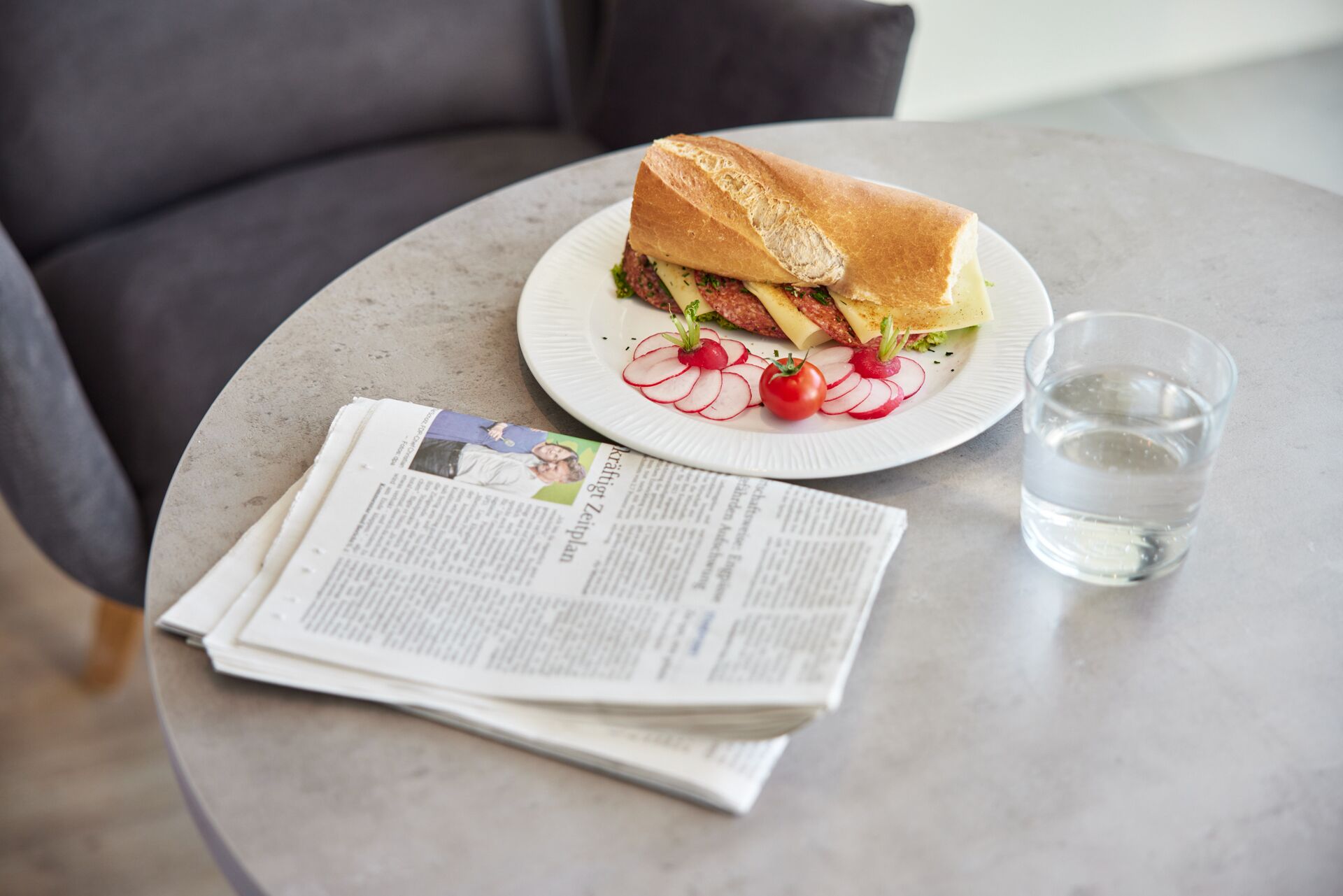 Un sándwich en un plato con un vaso de agua y un periódico sobre una mesa redonda.