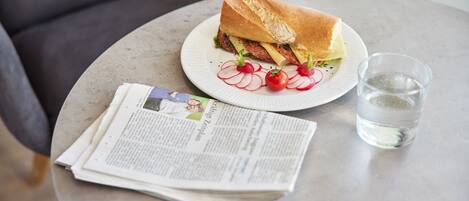 Un sándwich en un plato con un vaso de agua y un periódico sobre una mesa redonda.