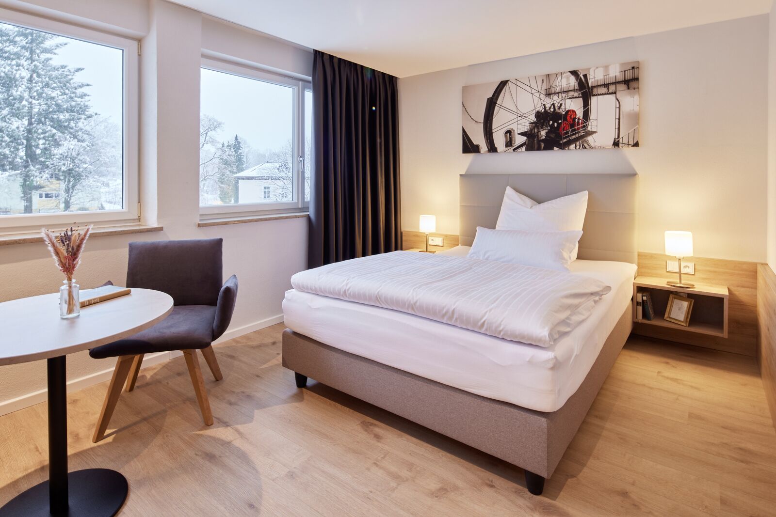 Une chambre d’hôtel moderne avec un grand lit, un bureau avec une chaise, une fenêtre donnant sur des arbres couverts de neige et une œuvre d’art accrochée au mur.