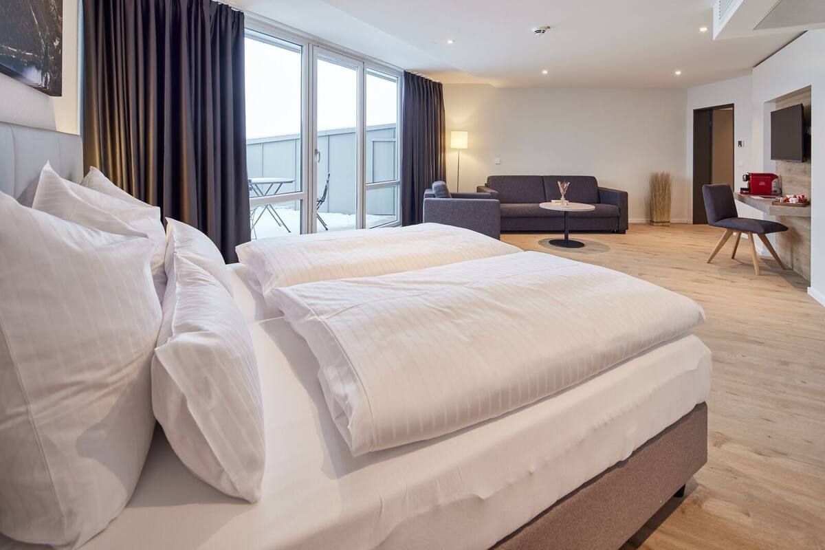 A modern hotel room with a large bed, a sofa, a desk, and a view of the outdoors.