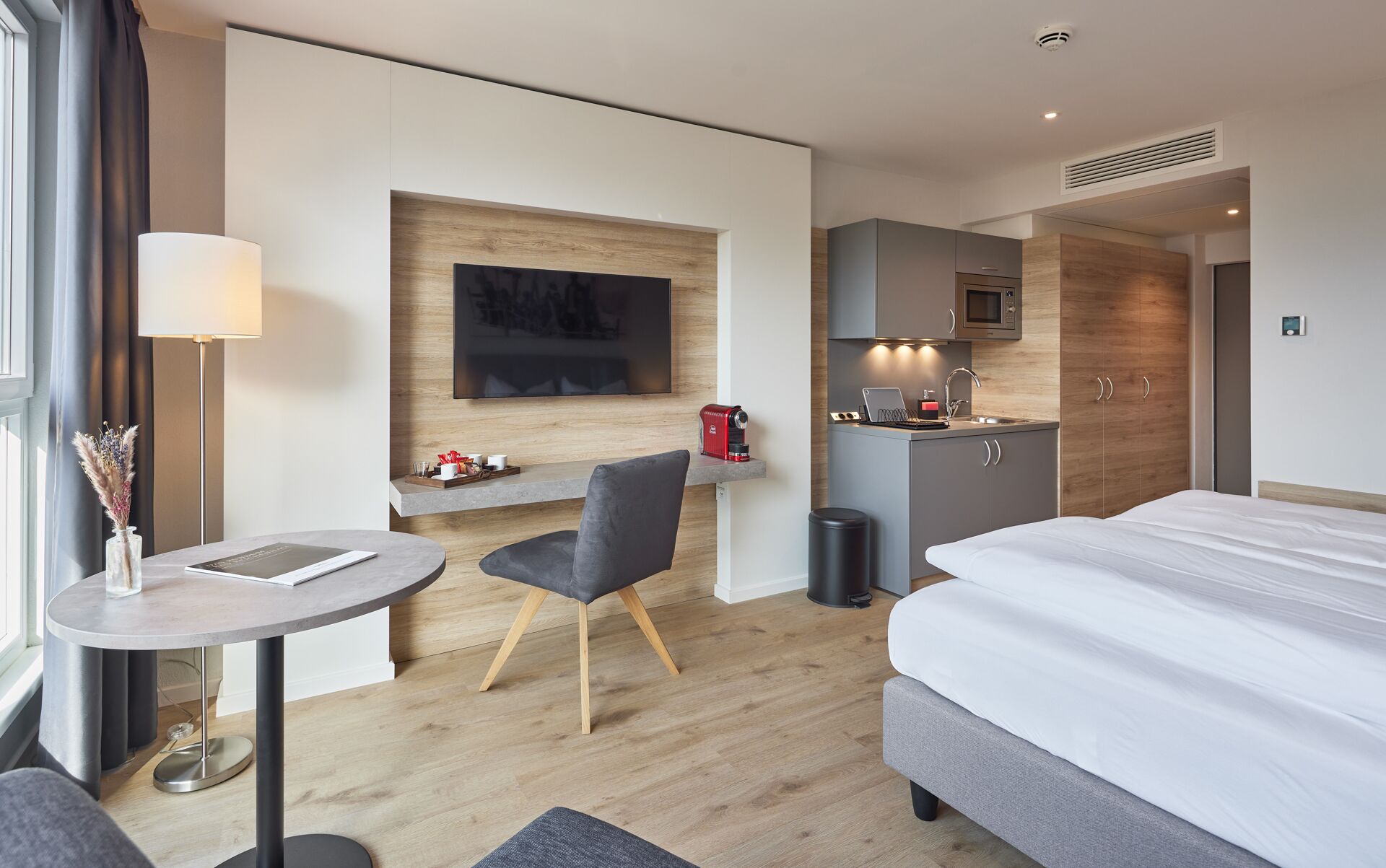 A modern hotel room with a bed, a desk with a lamp, a chair, a small kitchenette, and a TV mounted on the wall.