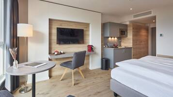 A modern hotel room with a bed, a desk with a lamp, a chair, a small kitchenette, and a TV mounted on the wall.