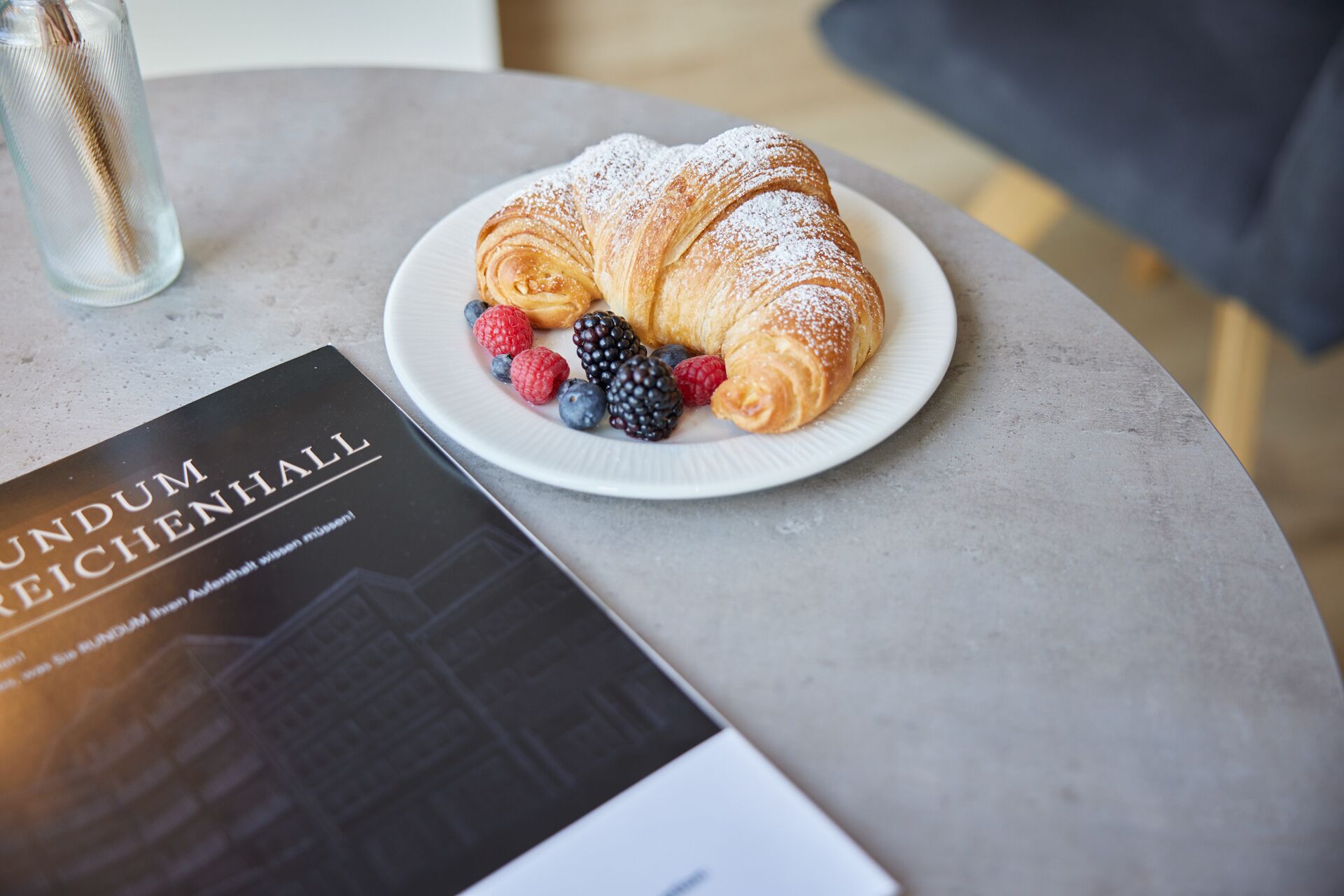 En croissant på en tallrik med bär, ett glas vatten och en bok med titeln ”RUNDUM REICENHAHL”.