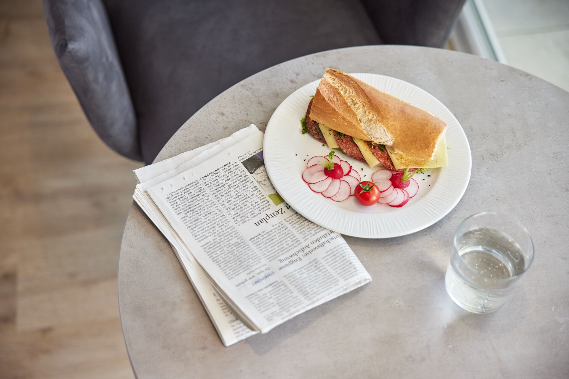 A sandwich on a plate with a glass of water and a newspaper on a round table.