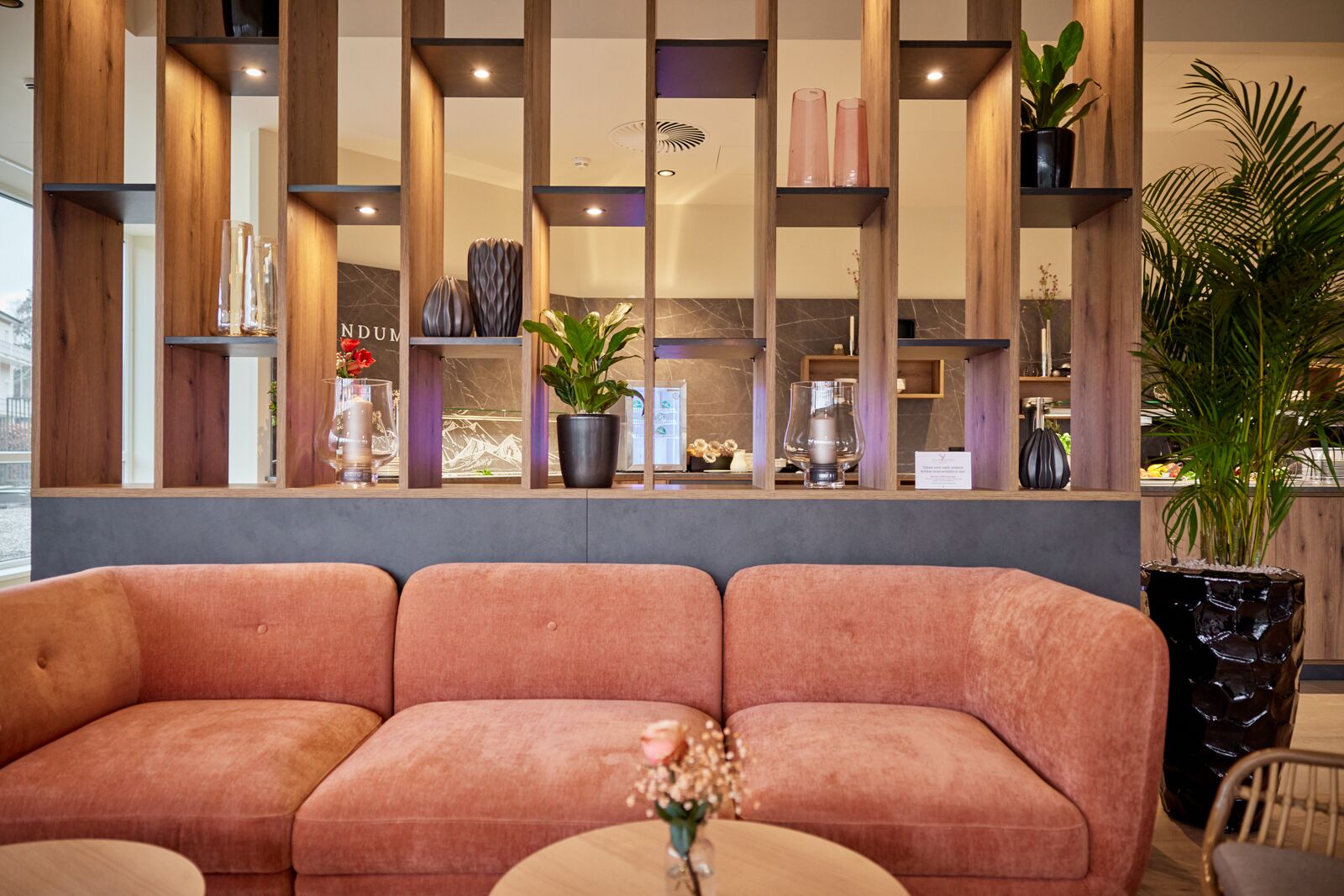 Una moderna área de recepción con un sofá rosa y mullido, un mostrador de madera y diversos objetos decorativos, como plantas y jarrones.