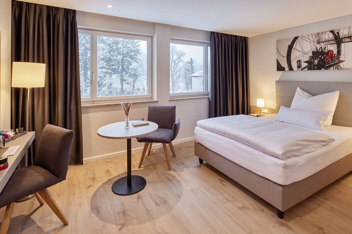 A hotel room with a bed, a desk, a chair, a round table, and a painting on the wall.