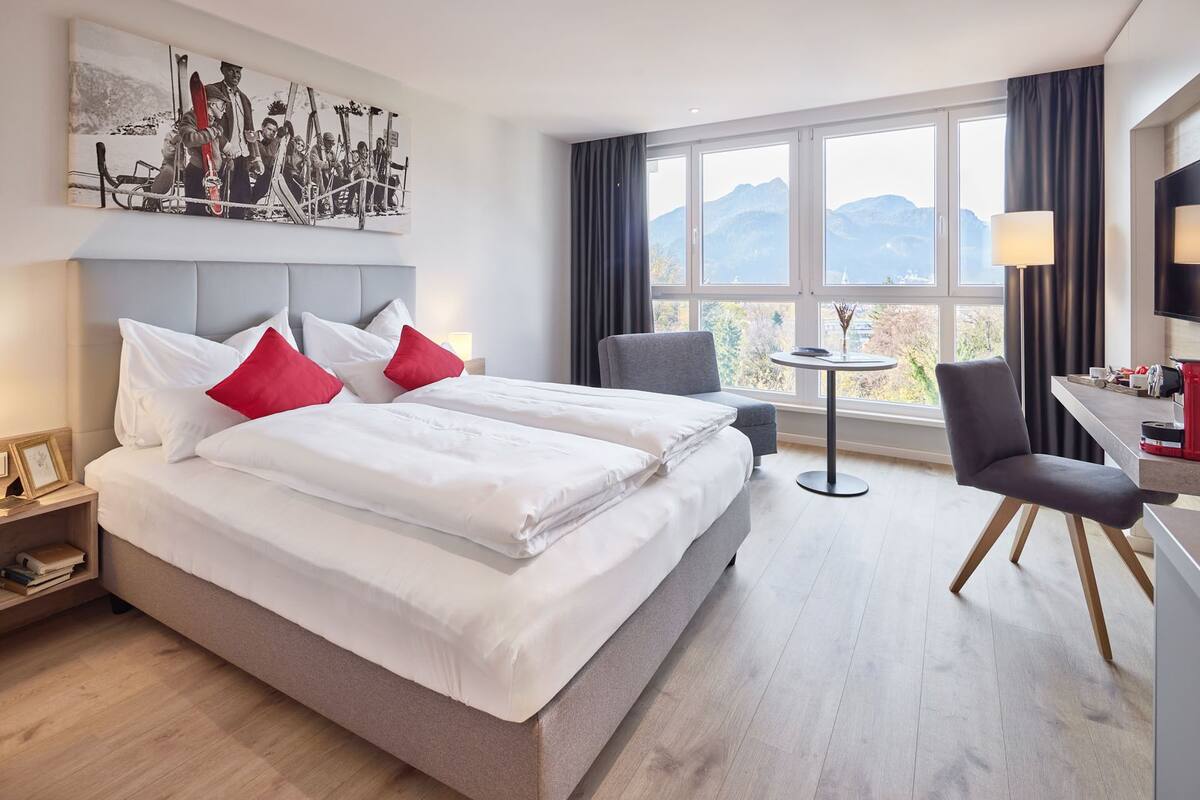 A hotel room with two beds, a small table, a chair, and a large window with a mountain view.