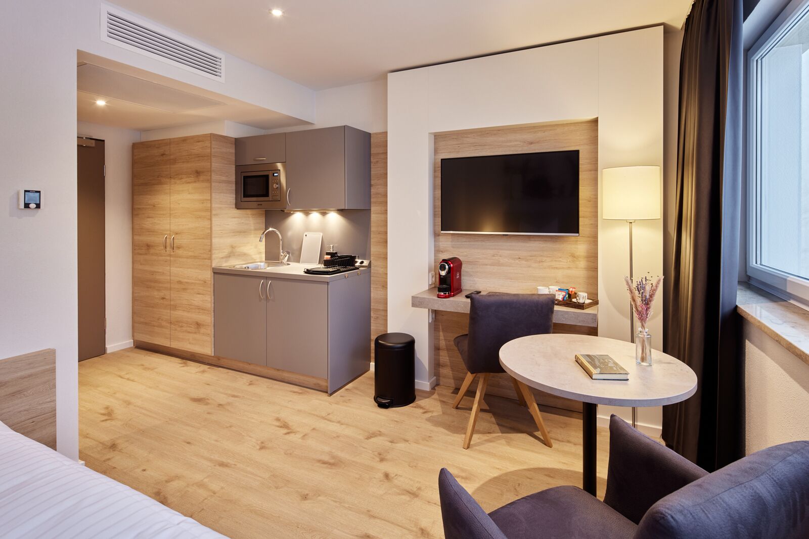 A modern hotel room with a kitchenette, a flat-screen TV, a round dining table, and a sofa.