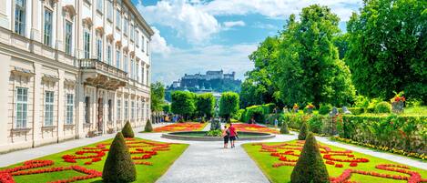 Un jardín formal con parterres simétricos, un camino central y un edificio grande de arquitectura clásica.