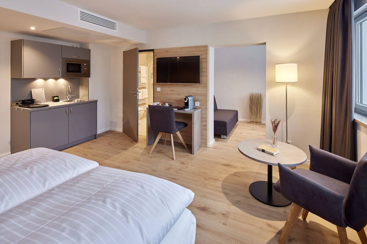 A hotel room with a bed, a desk with a chair, a TV, a microwave, and a small table with a lamp.