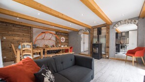 Una sala de estar moderna con un sofá gris, vigas de madera, una zona de comedor con mesa y sillas, y una cocina visible a través de un arco.
