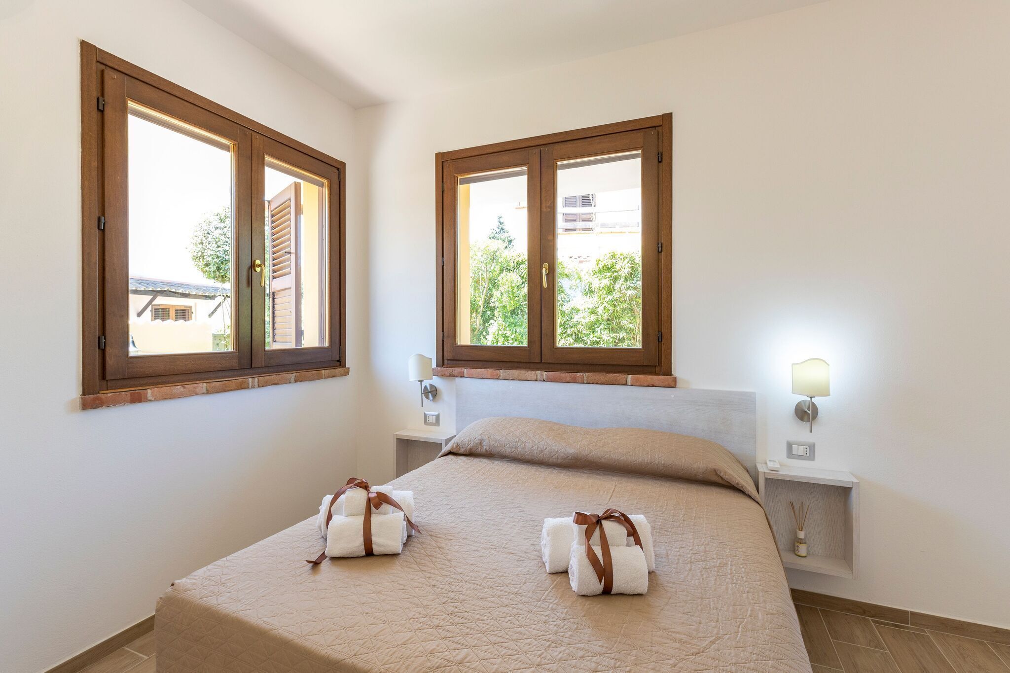 Une chambre d’hôtel comprenant un lit, deux tables de chevet, une petite table et deux fenêtres avec des cadres en bois.